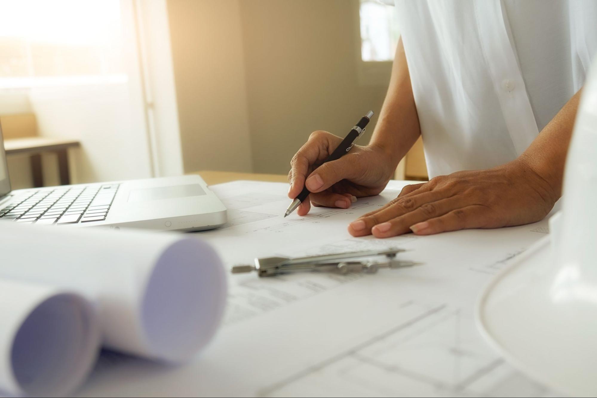 Uma engenheira projetando, em papel, com um computador sobre a mesa.